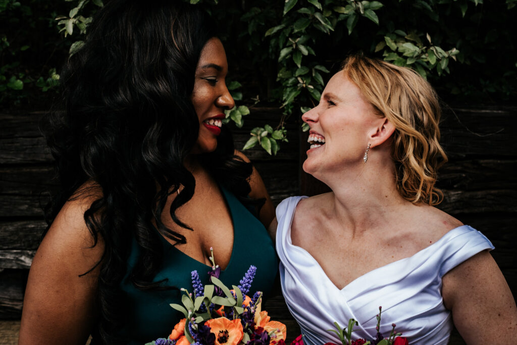 bride in white off the shoulder dress looking and laughing with her bridesmaid in a green dress and black hair