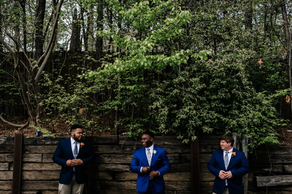 groom with his two groomsmen standing in a line all buttoning up their suit jackets looking at each other with greenery in the background