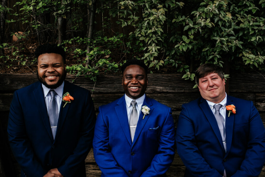 groom in navy suit with two groomsmen smiling at camera with greenery in the background