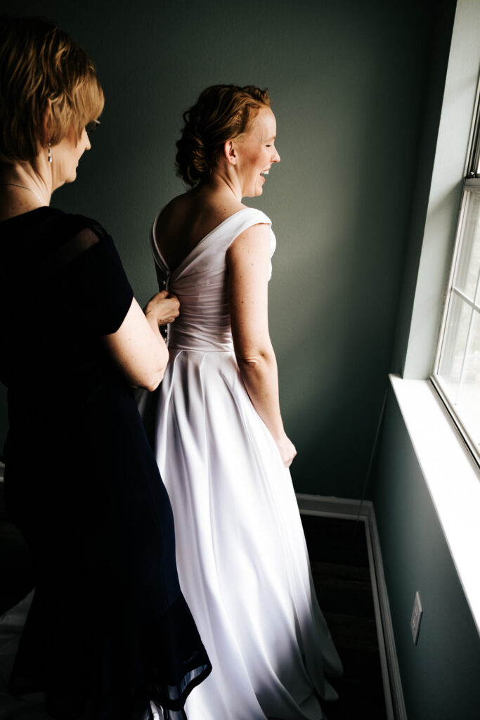 bride's mom in dark dress helping bride into her white wedding dress by zipping up the back while in front of a window