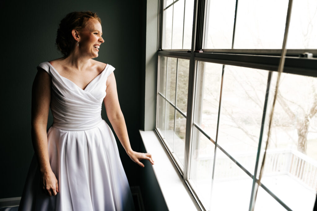 bride standing in front of window with hand on the window sill looking and smiling out the window