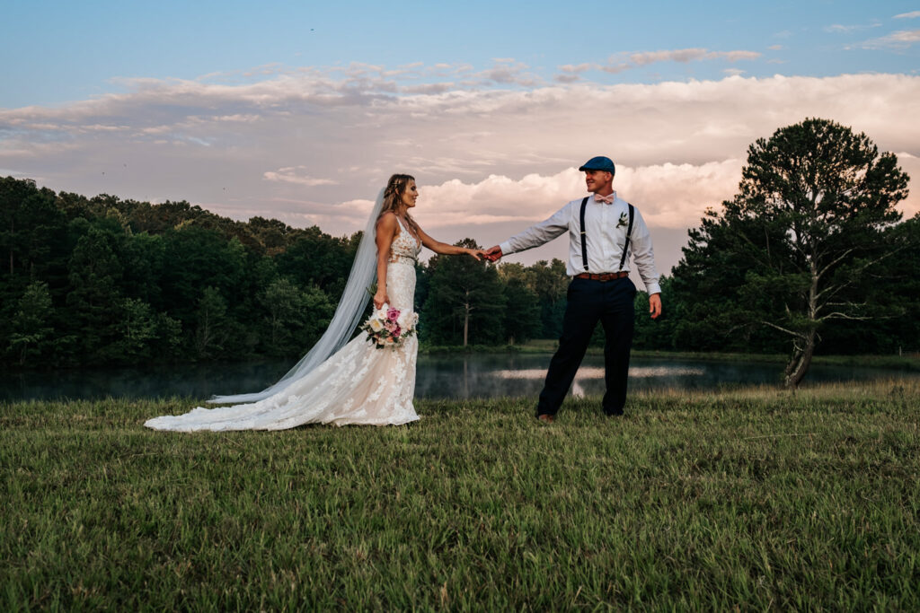 groom leading bride through the grass with blue and orange sky behind them