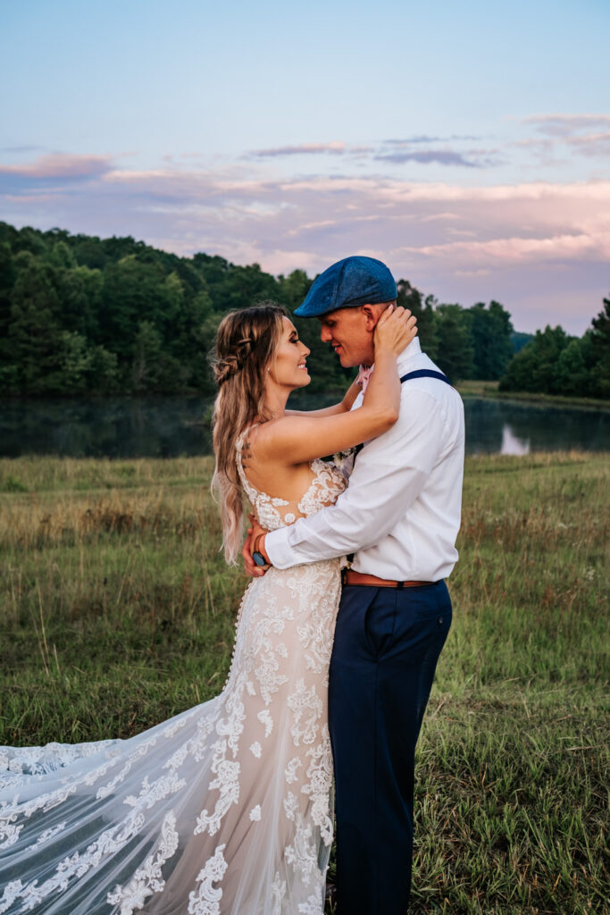 Bride with lace dress being held by groom with a blue hat with sunset in the background