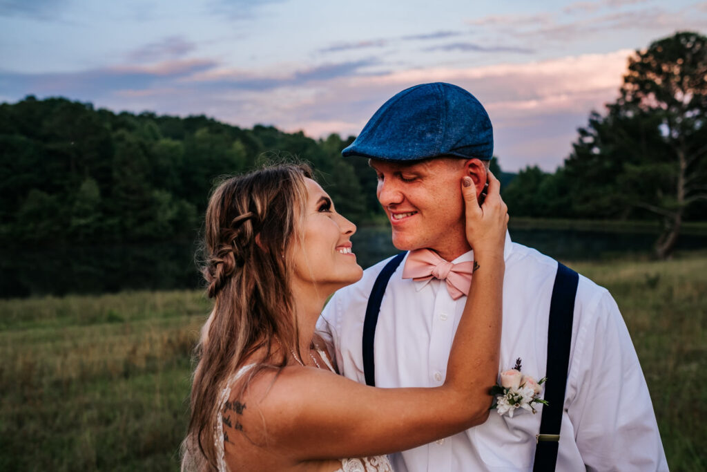 Bride holding the grooms jaw while they smile at each other with a sunset in background