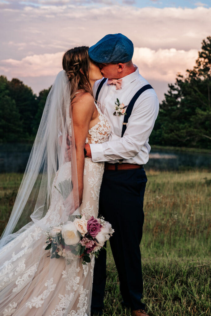 groom with blue hat puling bride in lace dress in for a kiss while she holds bouquet down