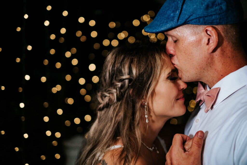 groom kissing brides forehead with hanging lights in the background