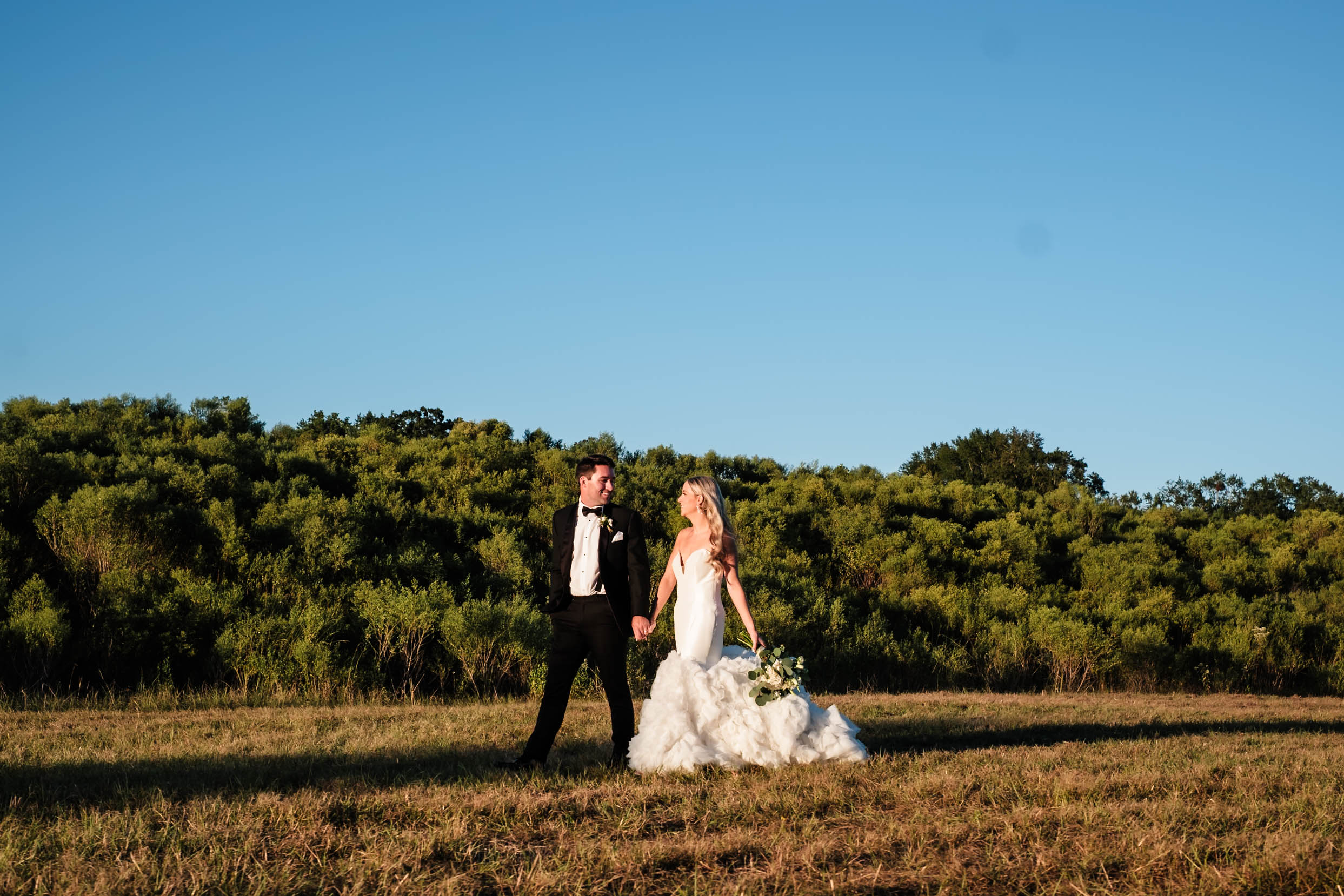 Bride and groom walking hand in hand across an open field at sunset, with lush greenery in the background. The bride wears a flowing white gown, and the groom is dressed in a classic black tuxedo, both sharing a joyful moment after planning a luxury wedding day.